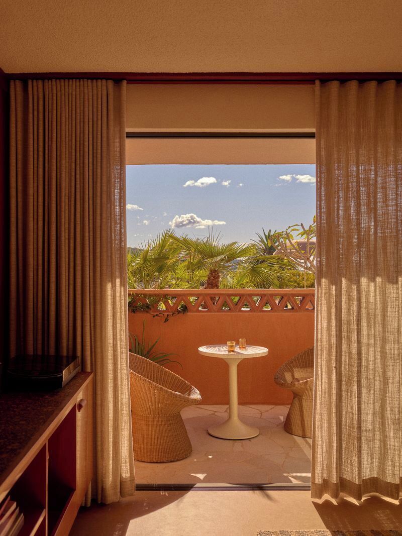 A room with a view of palm trees and a clear sky, a small round table, and two wicker chairs.