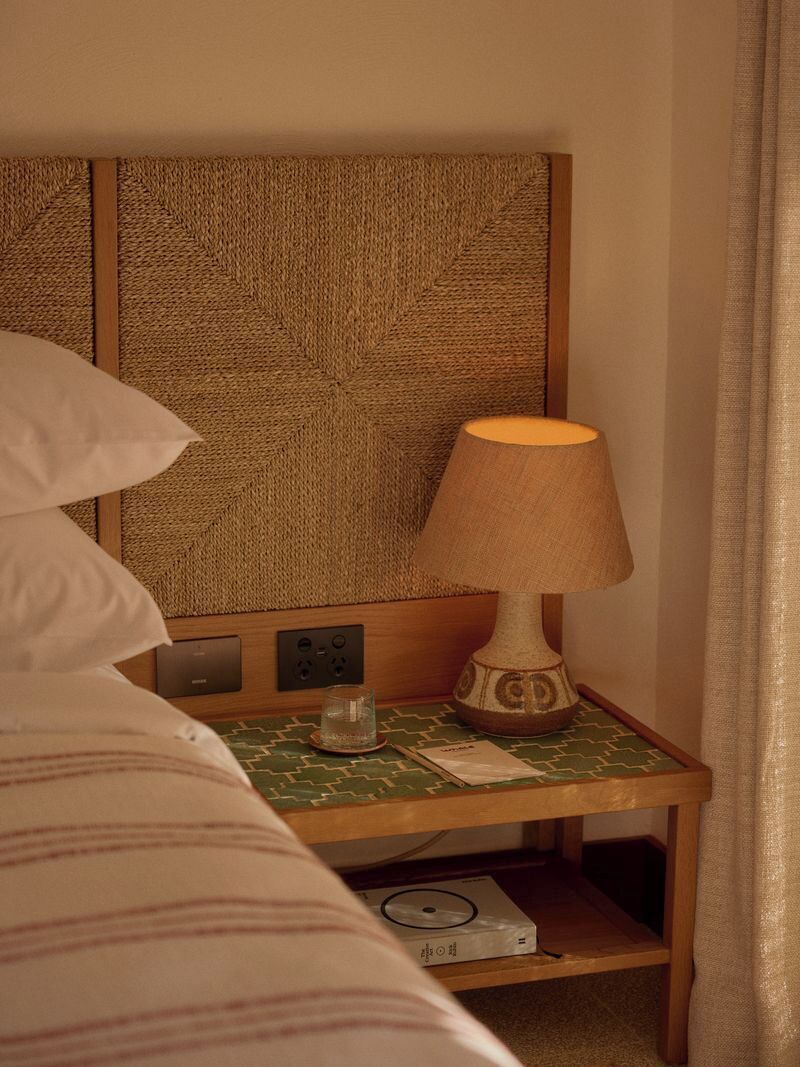 A hotel room with a bed, a nightstand with a lamp, a remote control, and a small shelf with a book.