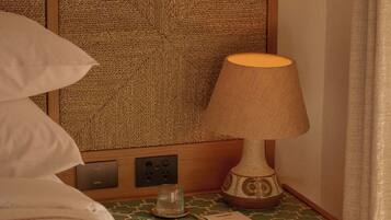 A hotel room with a bed, a nightstand with a lamp, a remote control, and a small shelf with a book.