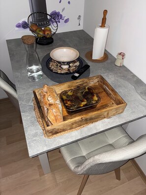 Une table Ă manger sur laquelle se trouve un plateau en bois, avec un plat rĂŽti, un bol de noix, une tĂ©lĂ©commande et un verre dâeau.