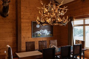 Een houten interieur met een eettafel, stoelen, een kroonluchter, een hertenkop en een berglandschap.