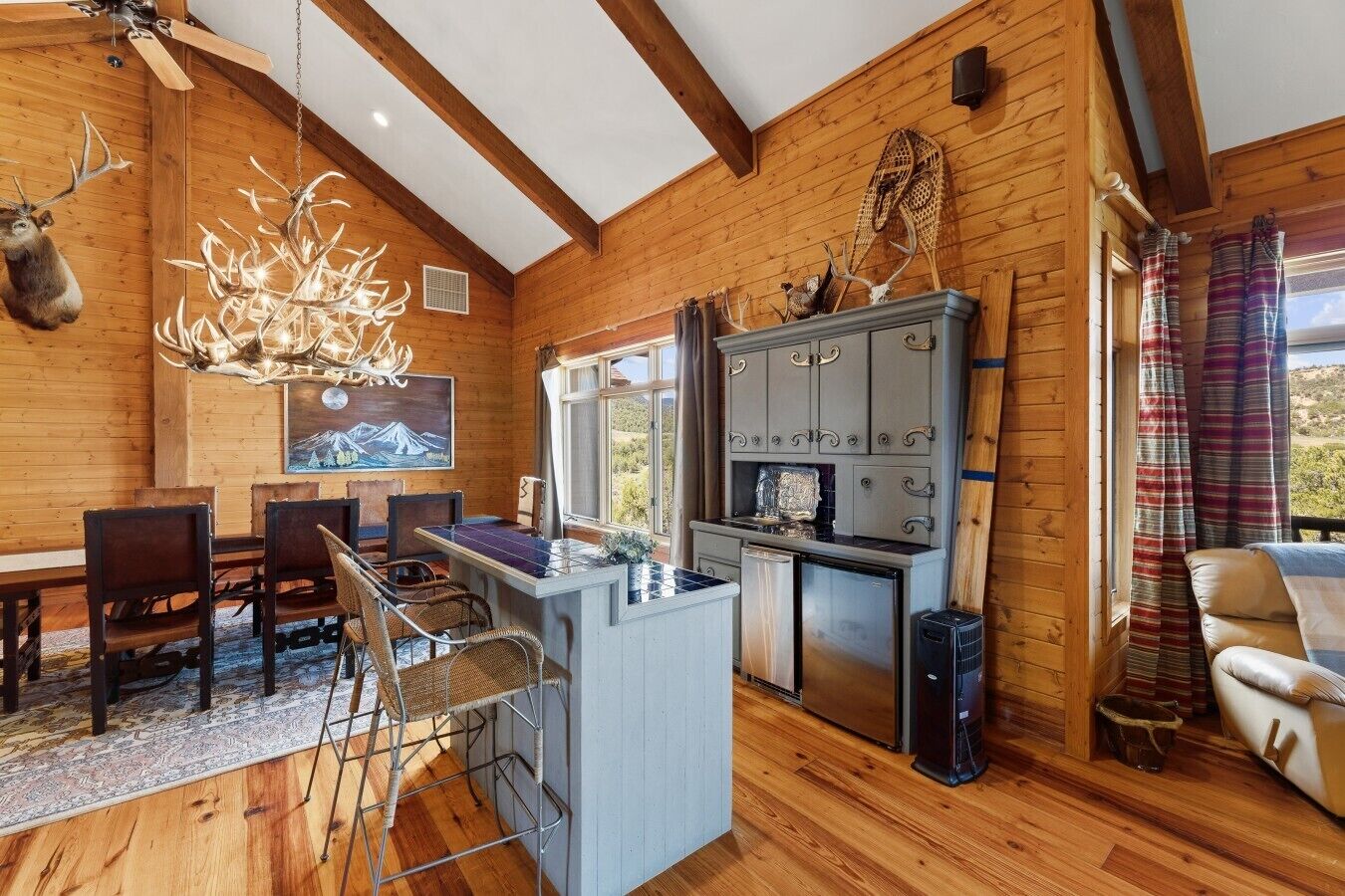 A wooden cabin interior with a dining area, kitchen, and a view of the outdoors.