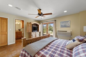 A bedroom with a bed, a ceiling fan, a window with curtains, a mounted TV, and a view of the outdoors.