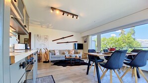 Une cuisine moderne dotée d’appareils électroménagers intégrés, un coin repas avec une table et des chaises en bois, un canapé et une grande fenêtre offrant une vue sur la verdure.