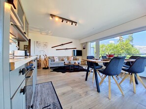 Eine moderne Küche mit integrierten Geräten, ein Essbereich mit Holztisch und -stühlen, eine Couch und ein großes Fenster mit Blick auf Grünflächen.