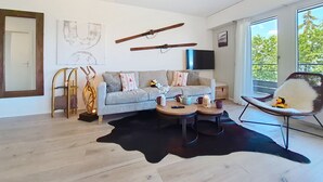 Un salon moderne avec un canapé, une table basse et un fauteuil. Une grande fenêtre et un téléviseur à écran plat fixé au mur complètent l’ensemble.