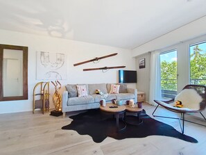 Ein modernes Wohnzimmer mit einer Couch, einem Couchtisch und einem Sessel. Ein großes Fenster und ein Flachbildfernseher sind an der Wand befestigt.