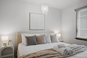 Een slaapkamer met een bed, twee nachtkastjes, een kroonluchter, een ingelijst schilderij en een raam.