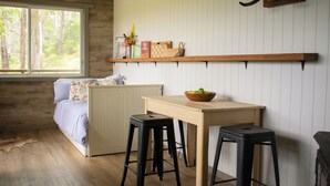 A room with a bed, a wooden table, two stools, a shelf with books and decorative items, and a window with a view of trees. - Tranquil secluded mountain chalet- perfect for relaxing! (Mount Byron)