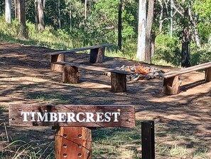 Un espace extĂ©rieur amĂ©nagĂ© dans un style rustique, avec un foyer, entourĂ© dâarbres. Une enseigne en bois portant lâinscription « TIMBERCREST » est visible.