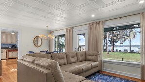 Ein Wohnzimmer mit einem großen, beigefarbenen Ecksofa, einem Essbereich mit Tisch und Stühlen, einer Küche mit Schränken und einer Kaffeemaschine sowie einem großen Fenster mit Blick auf einen See und Bäume.