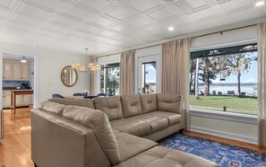 Una sala de estar con un amplio sofá seccional beige, un comedor con mesa y sillas, una cocina con armarios y cafetera, y un ventanal con vista a un lago y árboles.