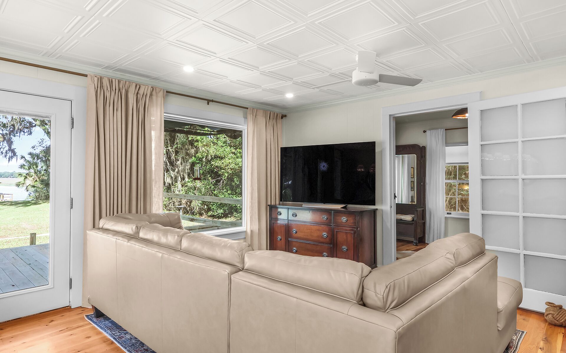 Un salon avec un canapé d’angle beige, un téléviseur posé sur un meuble en bois, un ventilateur de plafond et une vue sur l’extérieur.
