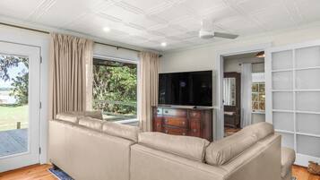 Un salon avec un canapé d’angle beige, un téléviseur posé sur un meuble en bois, un ventilateur de plafond et une vue sur l’extérieur.