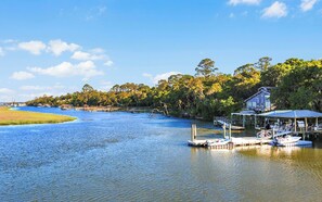 A riverside property with docked boats, a house with a covered porch, and a forested backdrop. - Waterfront Retreat on 2 Acres- Dock Access + Sunrise Views + Guest Suites (Townsend)