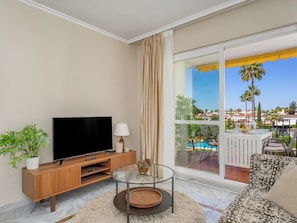 A living room with a TV, a glass coffee table, a sofa, and a view of a pool and palm trees. - San Pedro Beach by Interhome (Marbella)