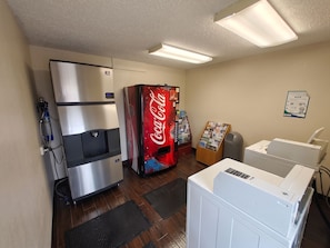Una habitación con refrigerador, máquina expendedora de Coca-Cola, un tablón de anuncios, un cubo de basura y una secadora.