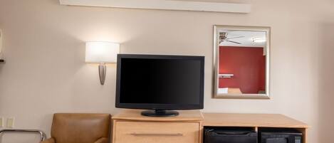 A hotel room with a television, a desk with drawers, a refrigerator, a microwave, a chair, and a wall-mounted lamp.