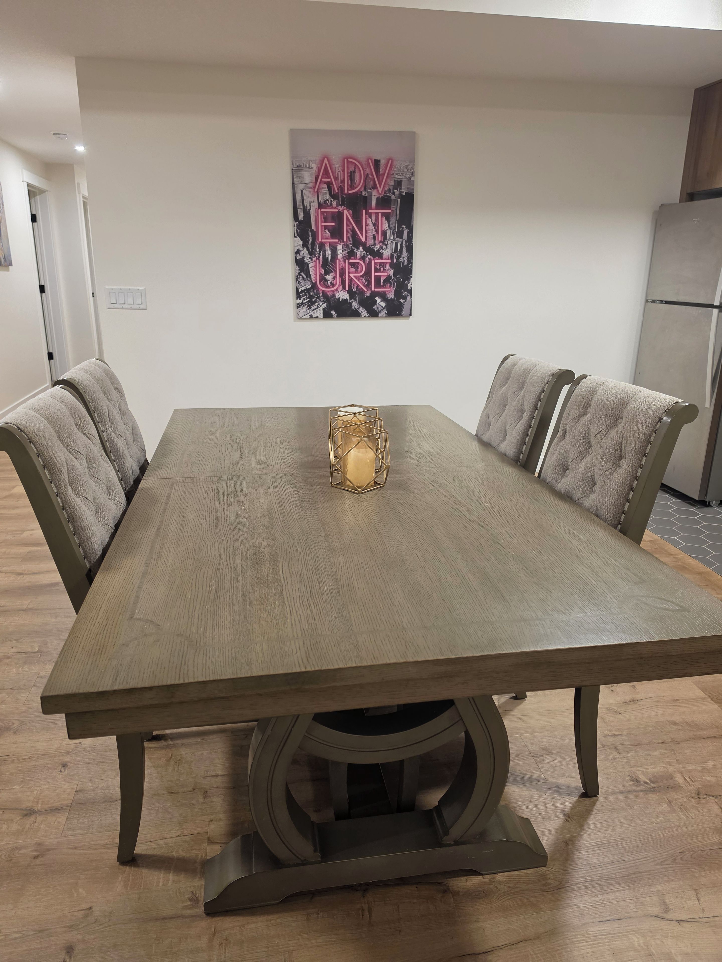 Une table de salle à manger rectangulaire, ornée d’un chandelier central, est entourée de chaises rembourrées à dossier capitonné. Une œuvre d’art murale portant le mot « AVENTURE » est visible au-dessus de la table.
