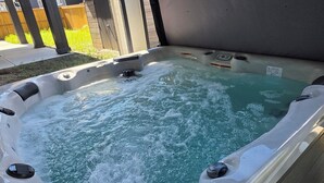 Un jacuzzi al aire libre con asiento integrado y panel de control, rodeado por una terraza de madera y una barandilla de metal.