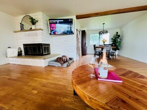 A living room with a fireplace, a TV mounted on the wall, a round wooden table with a bottle and glasses, and a dog resting on a cushion.