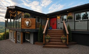 A modern two-story building with a wooden facade, a red door, and a balcony with string lights. - Okanagan Hideaway, Lake views & Outdoor Adventure, pet friendly and fun! (Vernon)