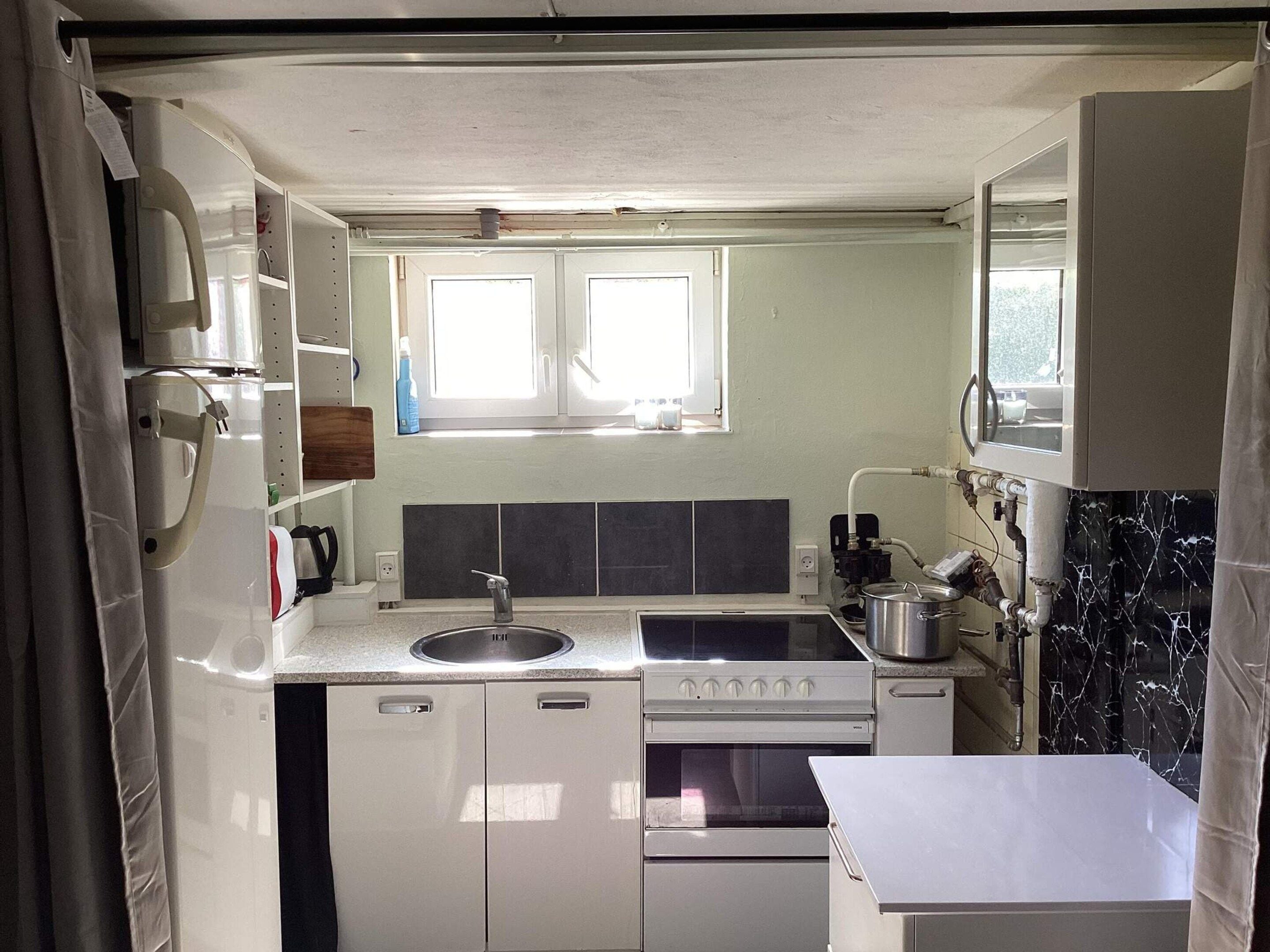 Een kleine keuken met een spoelbak, fornuis en oven, een raam boven de spoelbak, en een betegelde spatwand.