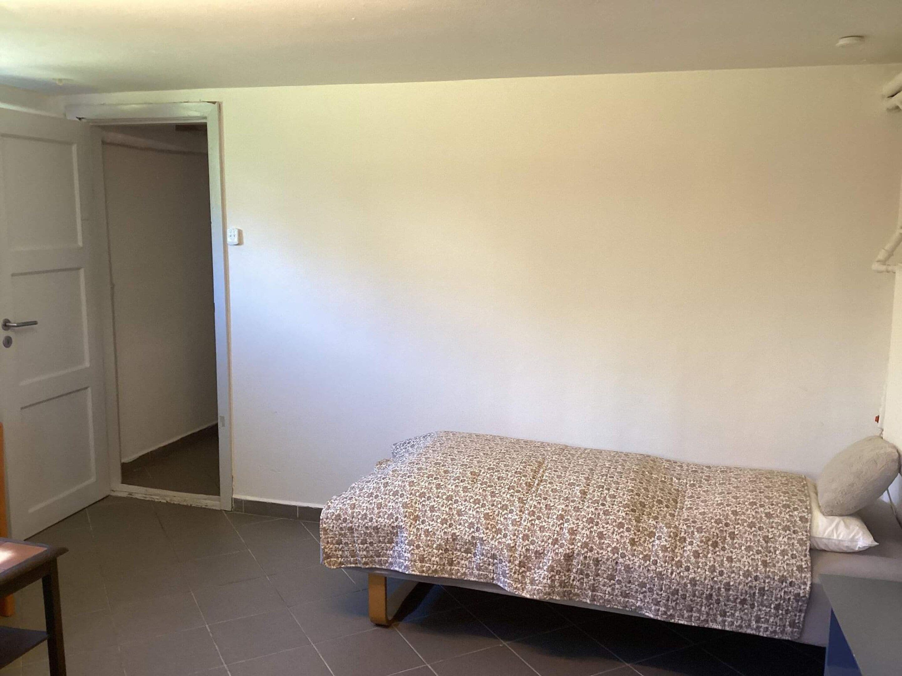 A small, single-bed room with a plain wall, a door, and a small table.