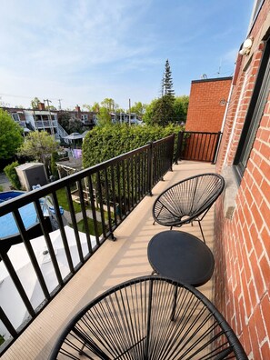 En balkon med to stole og et bord, en pool samt udsigt til omkringliggende bygninger og grønt.