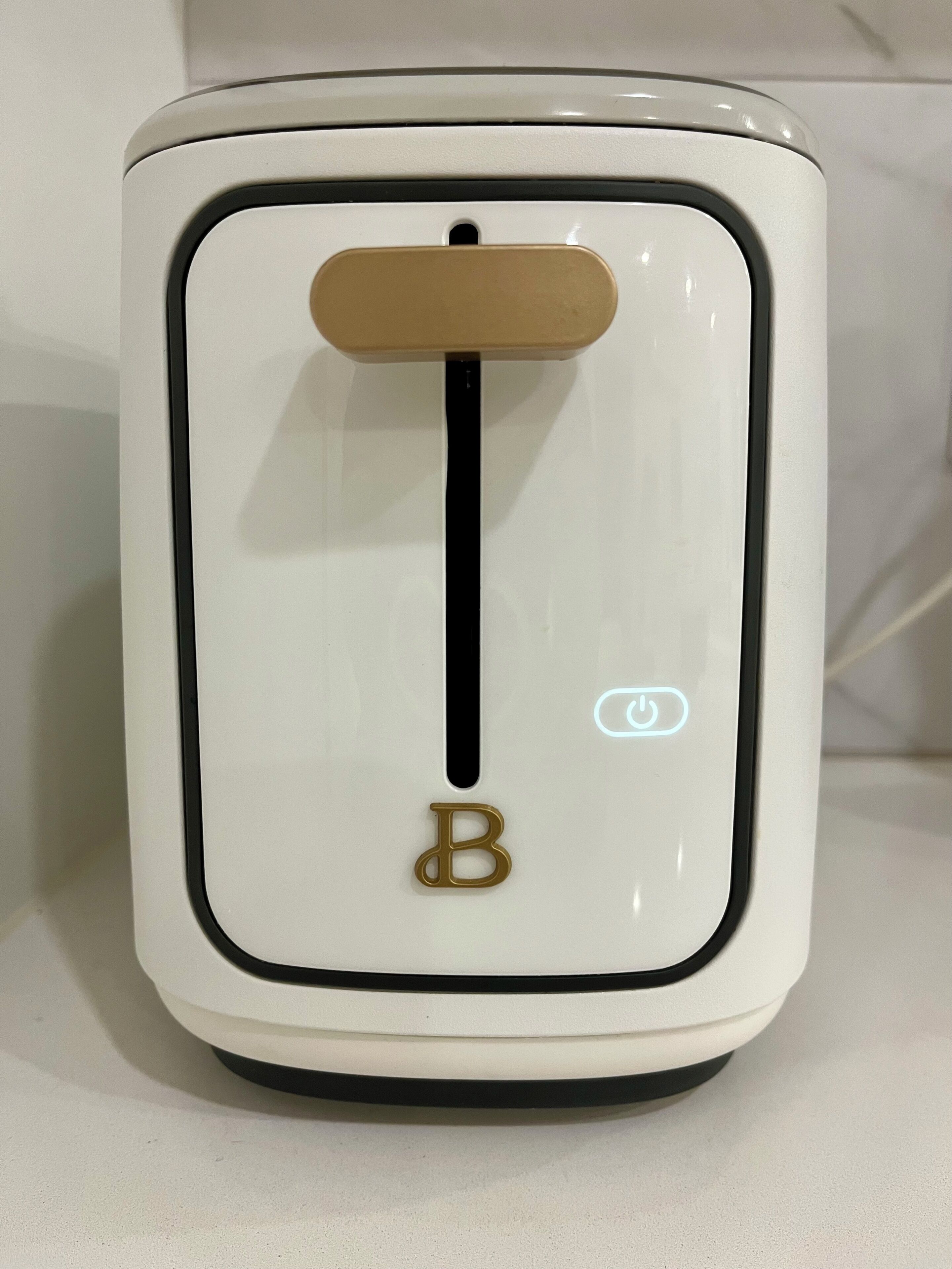 A white appliance with a black control panel, a gold letter 'B' button, and a power indicator light.