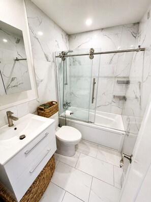 Et moderne badeværelse med glasvæg omkring brusebad, hvid vask og toilet.