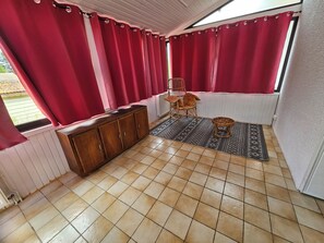 Una habitación con suelo de baldosas, un armario de madera, una silla de mimbre, un tapiz tejido y cortinas rojas.