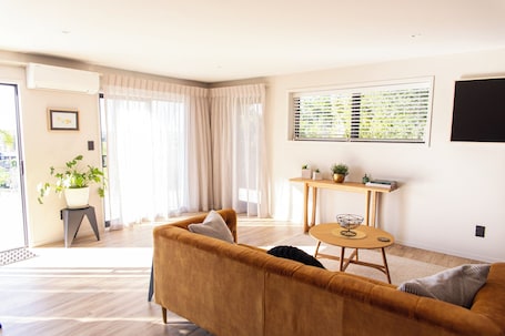 A living room with a sofa, a dining table with a bowl, a potted plant, and a window with blinds.. The Bee Hive