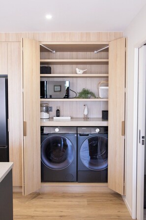 A compact laundry room with built-in washing machines, wooden shelving, and a small countertop.