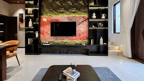 Een moderne woonkamer met een grote flatscreen televisie aan een gestructureerde wand, een donkere salontafel met een tijdschrift, en een boekenkast met decoratieve objecten.