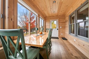 A wooden cabin interior with a dining table, chairs, and large windows offering a view of autumn trees.