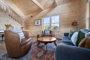 A cozy cabin interior with a wooden staircase, a large window, a round wooden coffee table, and a blue sofa.