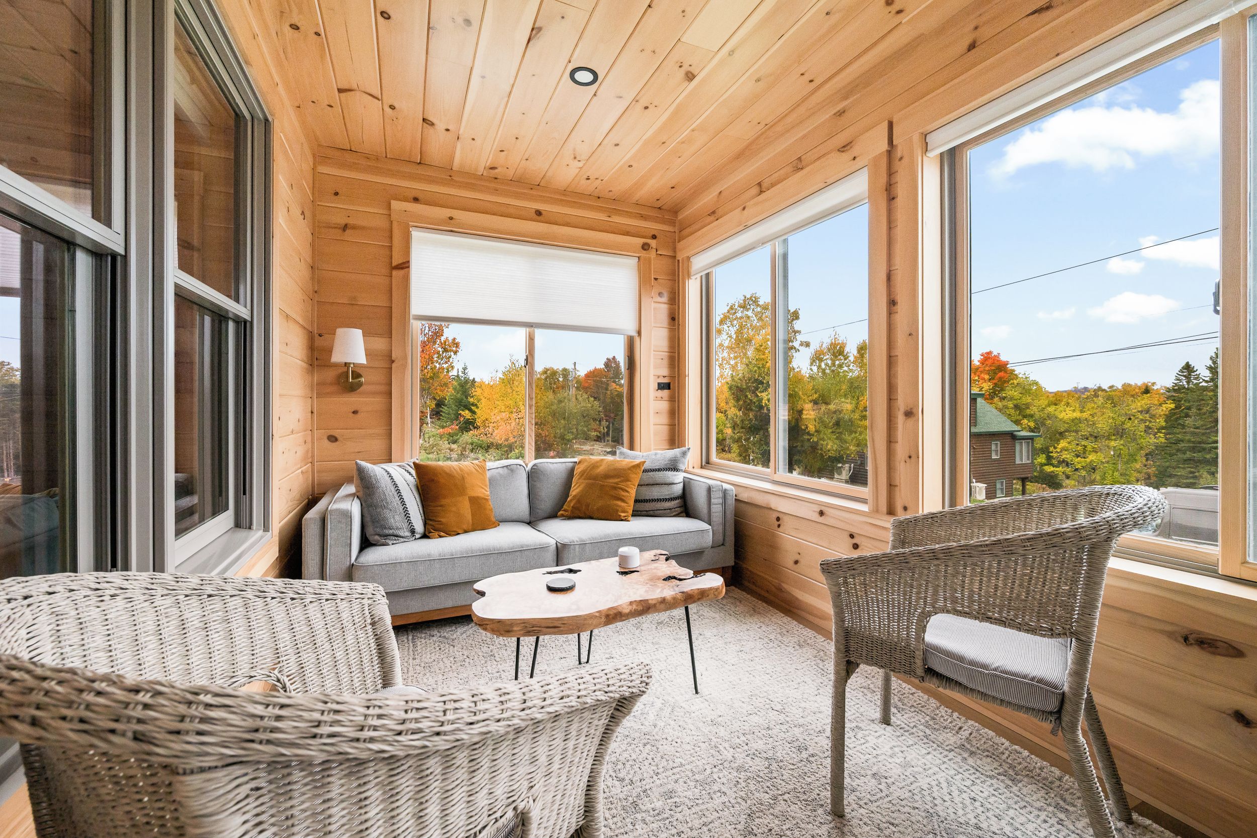 A cozy cabin interior with a sofa, two wicker chairs, a coffee table, and large windows offering a view of autumn trees.