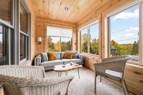 A cozy cabin interior with a sofa, two wicker chairs, a coffee table, and large windows offering a view of autumn trees.