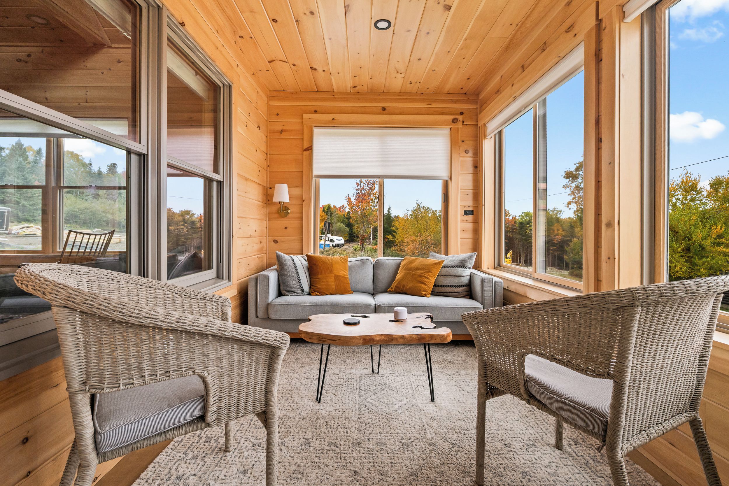 L’intérieur chaleureux d’un chalet avec un canapé, deux fauteuils en osier, une table basse et de grandes fenêtres offrant une vue sur les arbres et un ciel dégagé.
