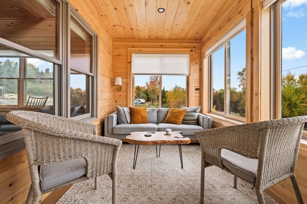 A cozy cabin interior with a sofa, two wicker chairs, a coffee table, and large windows offering a view of trees and a clear sky.