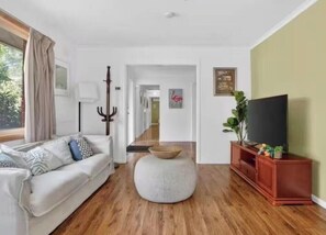A living room with a white sofa, a round ottoman, a wooden floor, a television on a wooden stand, and a plant. - 4B cozy holiday house by frankston sea (Frankston)