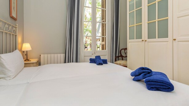 A neatly made bed with white linens, a bedside lamp, and blue towels on a white headboard. - Poble Nou - Llacuna by Interhome (Barcelona)