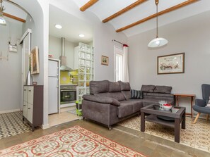 Ein Wohnzimmer mit grauem Sofa, einem kleinen Tisch und einem Sessel. Im Hintergrund befindet sich eine Küche mit Herd und Schränken.