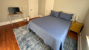 Un dormitorio con cama, escritorio con computadora, dos puertas y mesita de noche con lámpara.