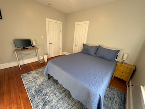 Un dormitorio con cama, escritorio con computadora, dos puertas y mesita de noche con lámpara.