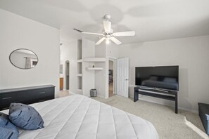 Un dormitorio con cama, televisión, ventilador de techo, espejo y puerta que da a otra habitación.