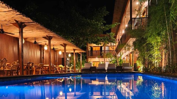 A nighttime pool area with lounge chairs and umbrellas, surrounded by buildings with balconies and vegetation. - Home Dejavu (Gili Trawangan)
