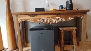 A compact GEA refrigerator under a wooden table with a decorative carving, a black coffee maker, and a clear water bottle on the table. - Home Dejavu (Gili Trawangan)