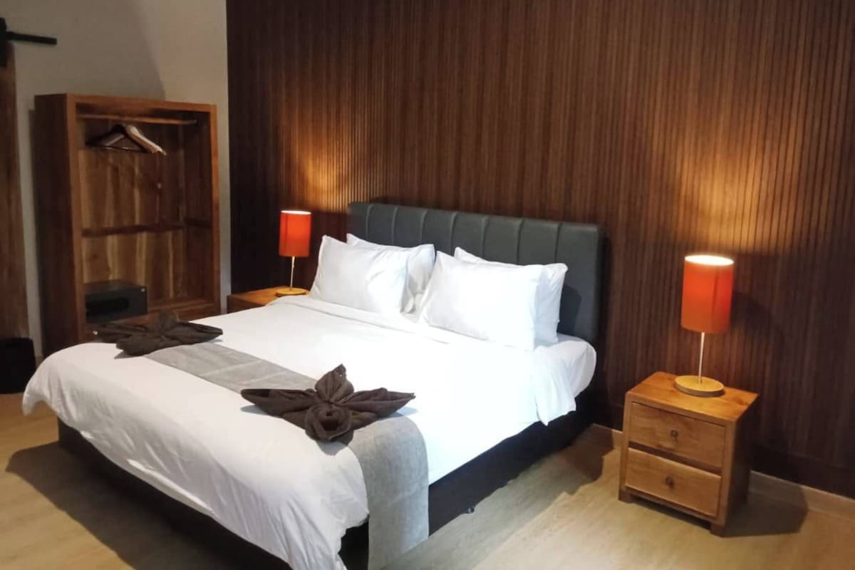 Una cama bien hecha con sábanas blancas, dos lámparas de noche, una mesita de noche de madera y un armario de madera.